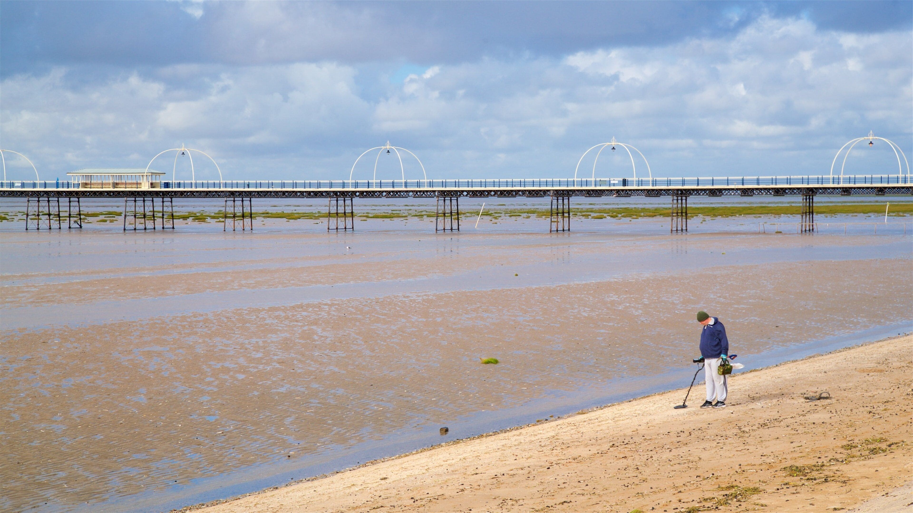 A beach with a person standing near the water's edge, a bridge in the background, and a cloudy sky.
