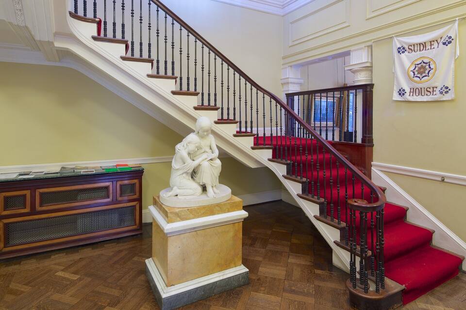 A grand staircase with red carpeted steps, a marble statue, and a banner reading "Sudley House".