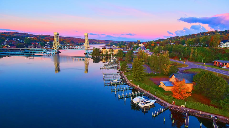 A calm river with a bridge, docked boats, and autumn-colored trees.