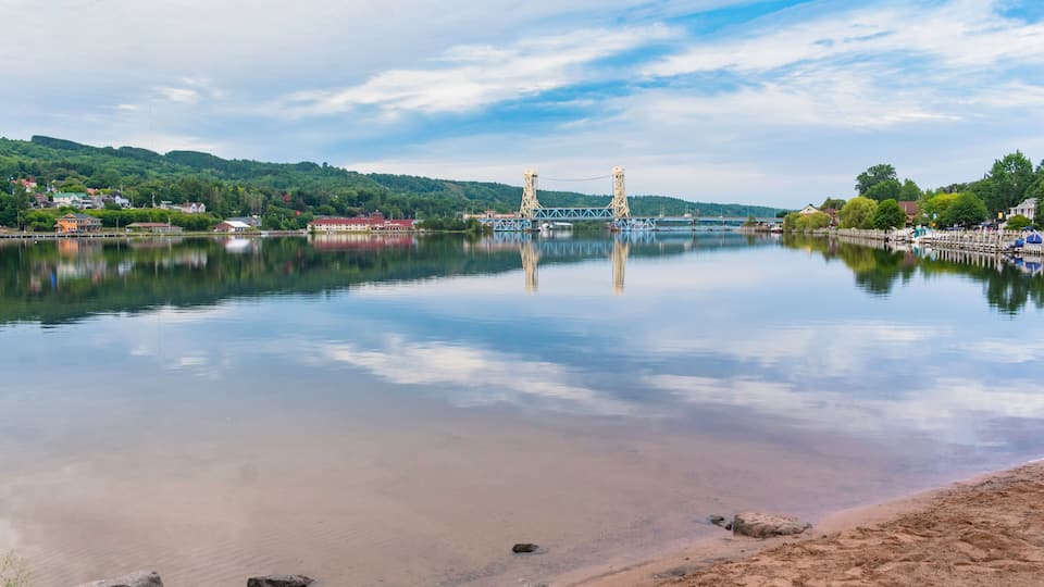 A calm river with a bridge, sandy shore, and green hills in the background.