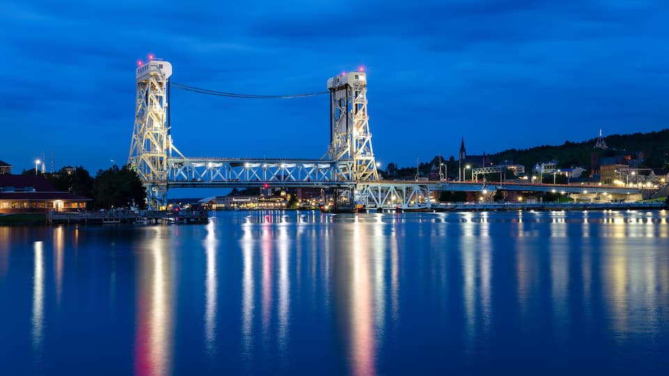 A large suspension bridge at night with its towers illuminated, spanning over a calm river with city lights in the background.