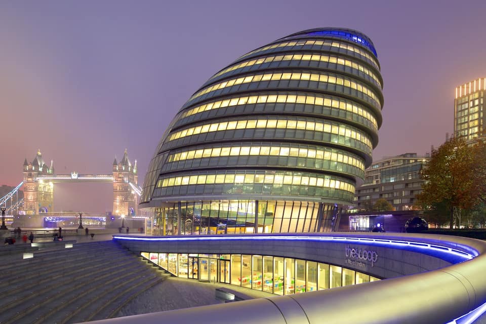 A modern curved building with illuminated windows, a bridge in the background, and a staircase leading to an upper level.