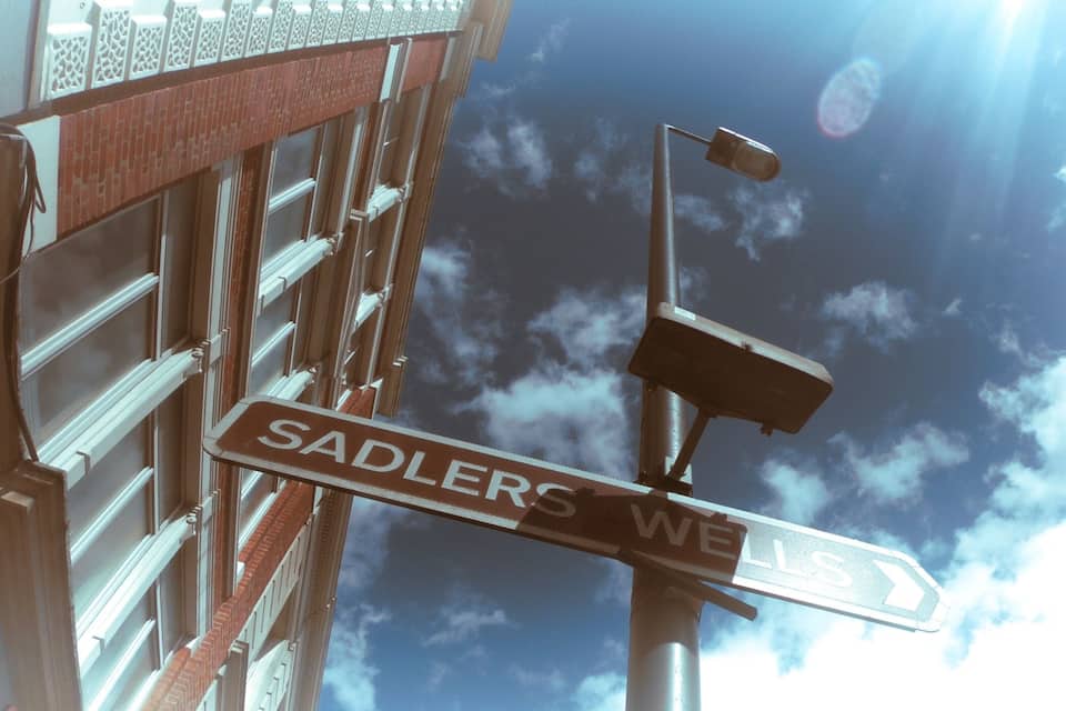 Una señal de calle para 'SADLER'S WELL' con un cielo azul claro y nubes al fondo.