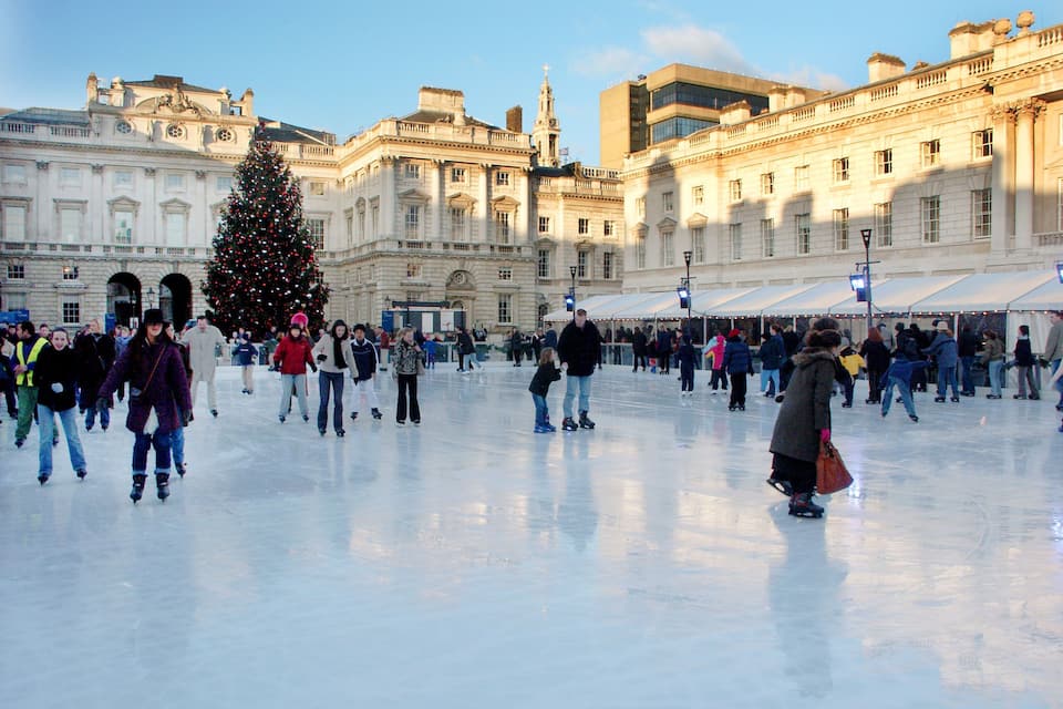 Uma grande pista de patinação ao ar livre rodeada por edifícios históricos, com uma árvore de Natal decorada no centro.