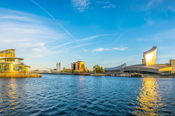 A waterfront cityscape with modern architecture, including a distinctive building with a curved roof and a cylindrical structure with a golden facade.