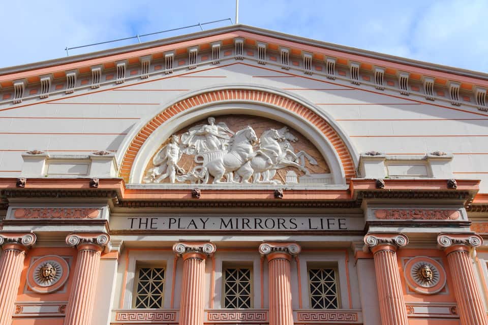 A building facade with columns, a central arch, and the inscription "THE PLAY MIRRORS LIFE".