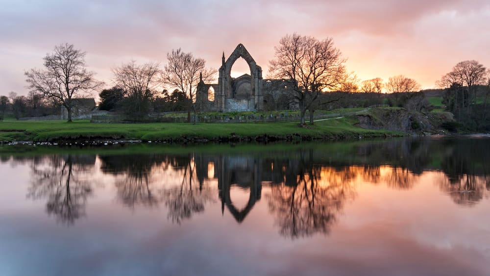 A historic stone structure with a pointed arch, surrounded by trees and a calm body of water reflecting the structure.
