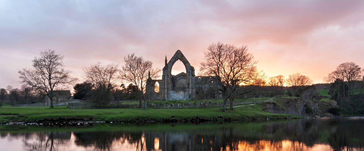 A historic stone structure with a pointed arch, surrounded by trees and a calm body of water reflecting the structure.