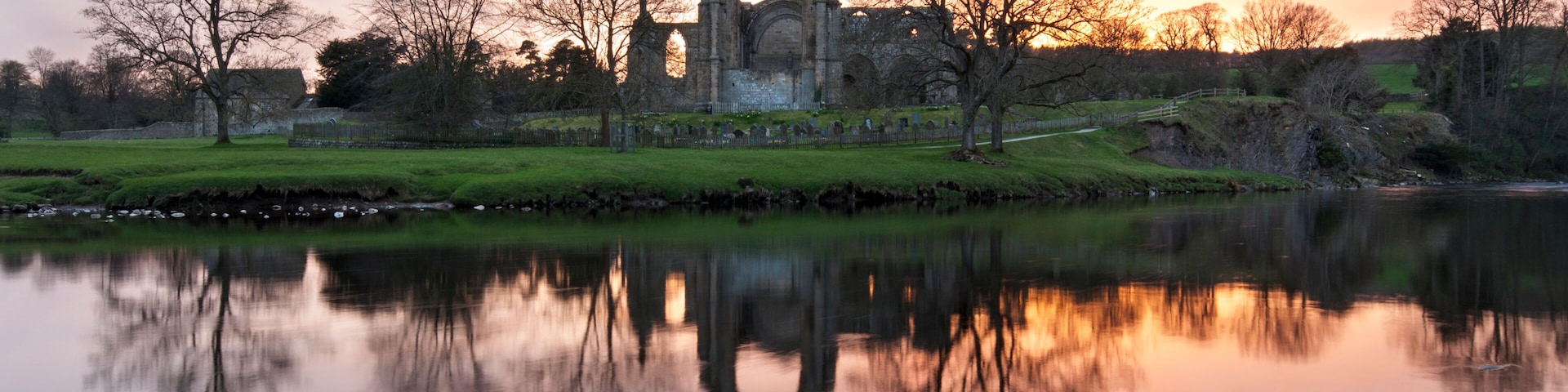 A historic stone structure with a pointed arch, surrounded by trees and a calm body of water reflecting the structure.