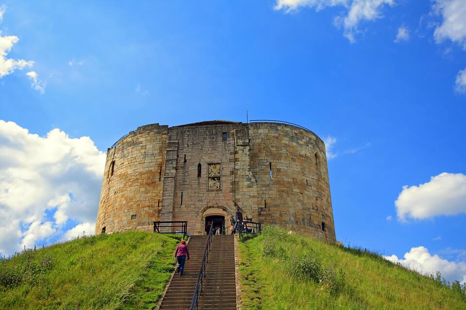 A stone tower with a staircase leading up to its entrance, surrounded by grass and under a blue sky with clouds.