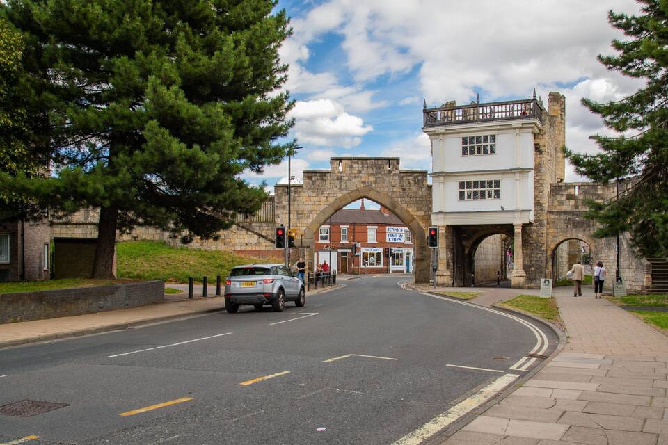 A street with a stone archway, a white building with a sign, and a car driving through.