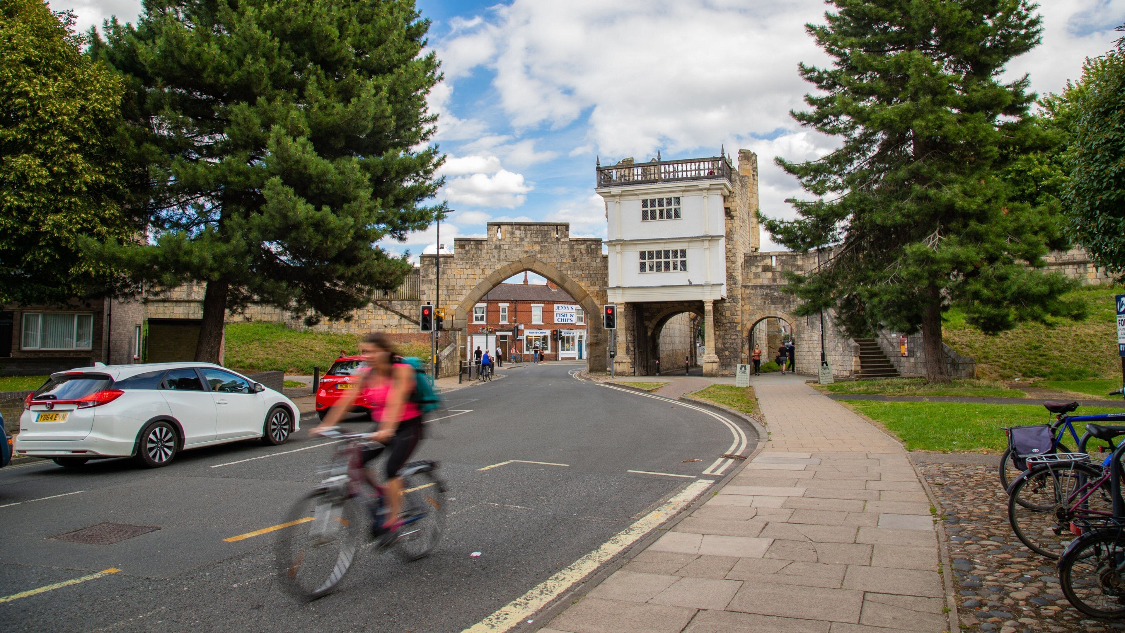 A street with a historic stone archway, a white building with a balcony, and bicycles parked on the sidewalk.
