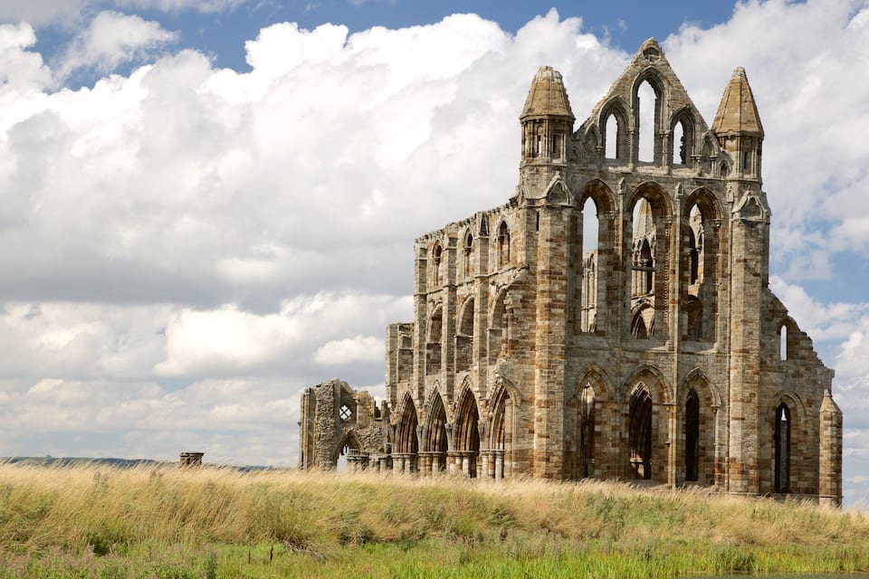 Une vaste cathédrale historique en pierre, dotée de nombreuses arcades et de tours, entourée de prairies verdoyantes et surplombée d’un ciel partiellement nuageux.