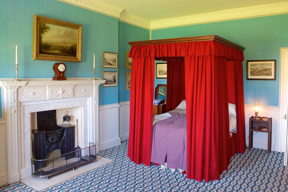 A bedroom with a four-poster bed, a fireplace, and patterned wallpaper.