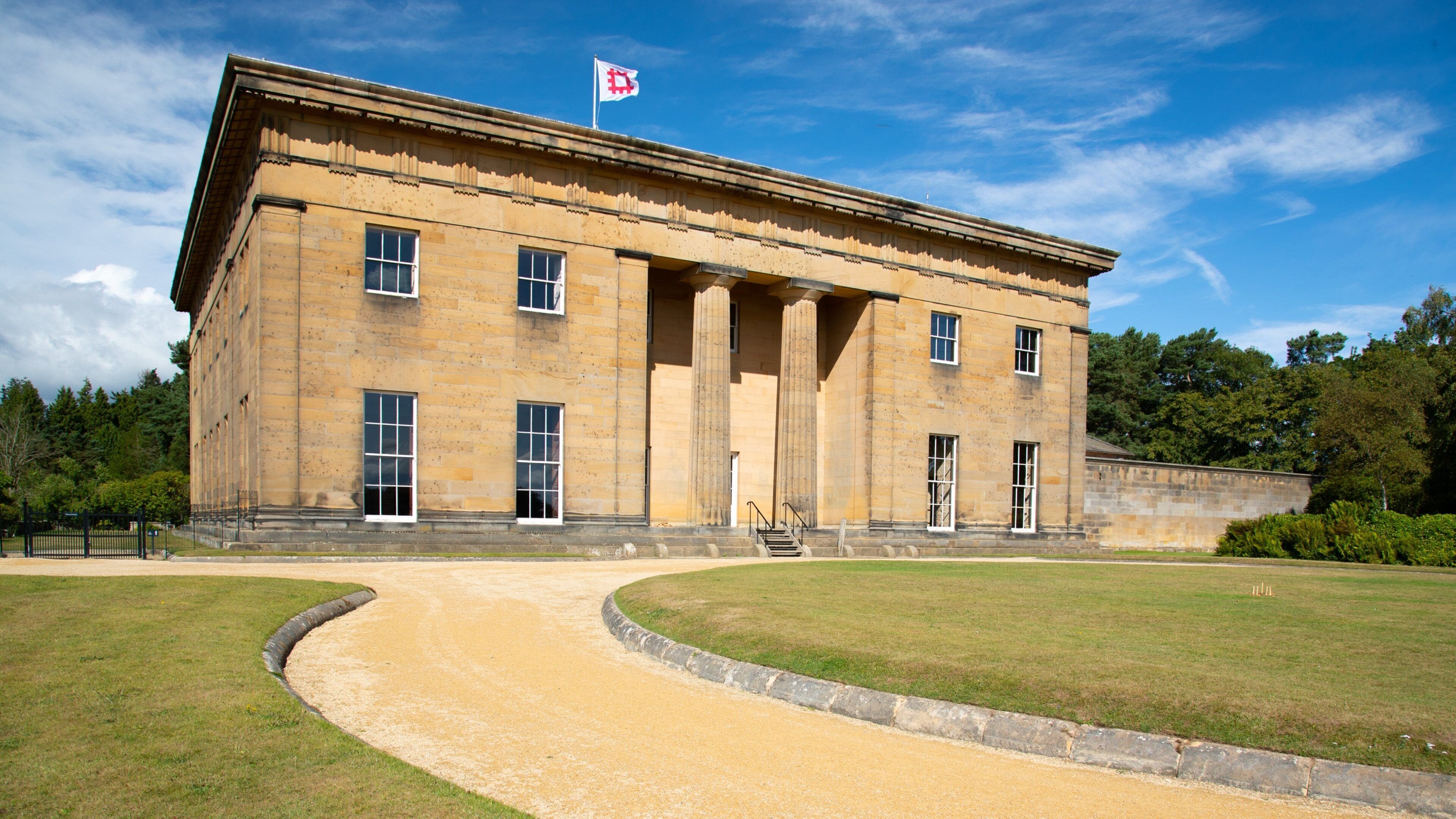 A large, historic building with a flag on top, surrounded by a well-maintained lawn and a curved pathway.