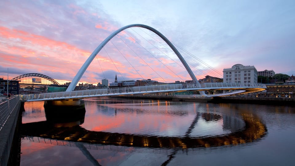 A modern bridge with a distinctive arch over a river, reflecting the sunset sky.