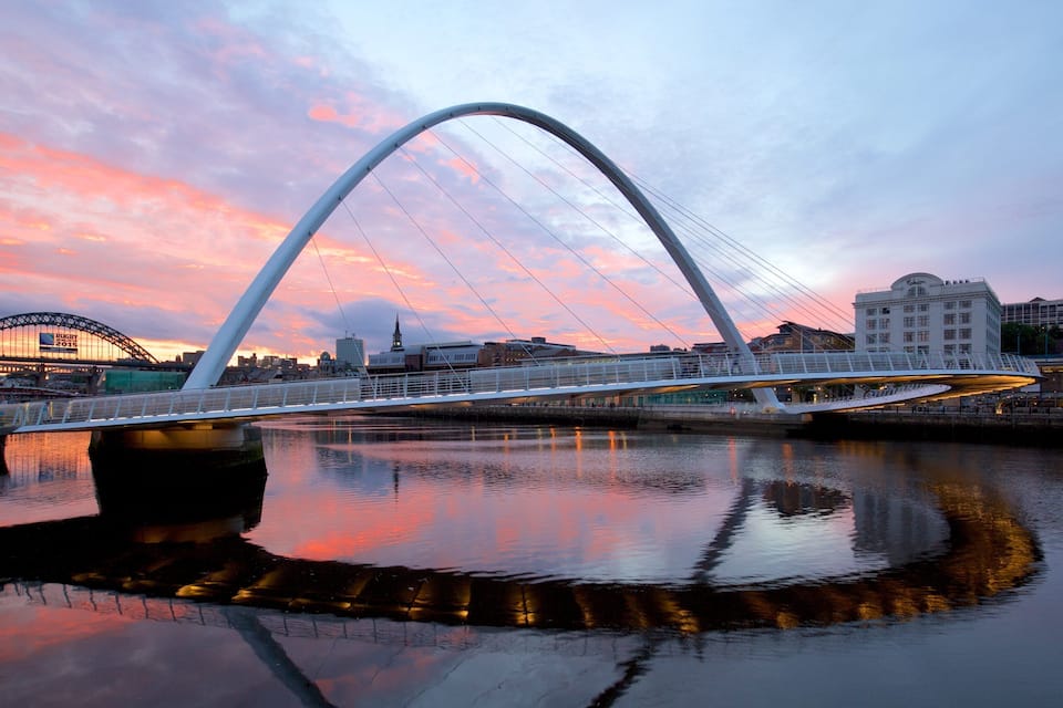 A modern bridge with a distinctive arch over a river, reflecting the sunset sky.