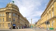 A historic street with classical architecture, a prominent tower, and a busy pedestrian area.