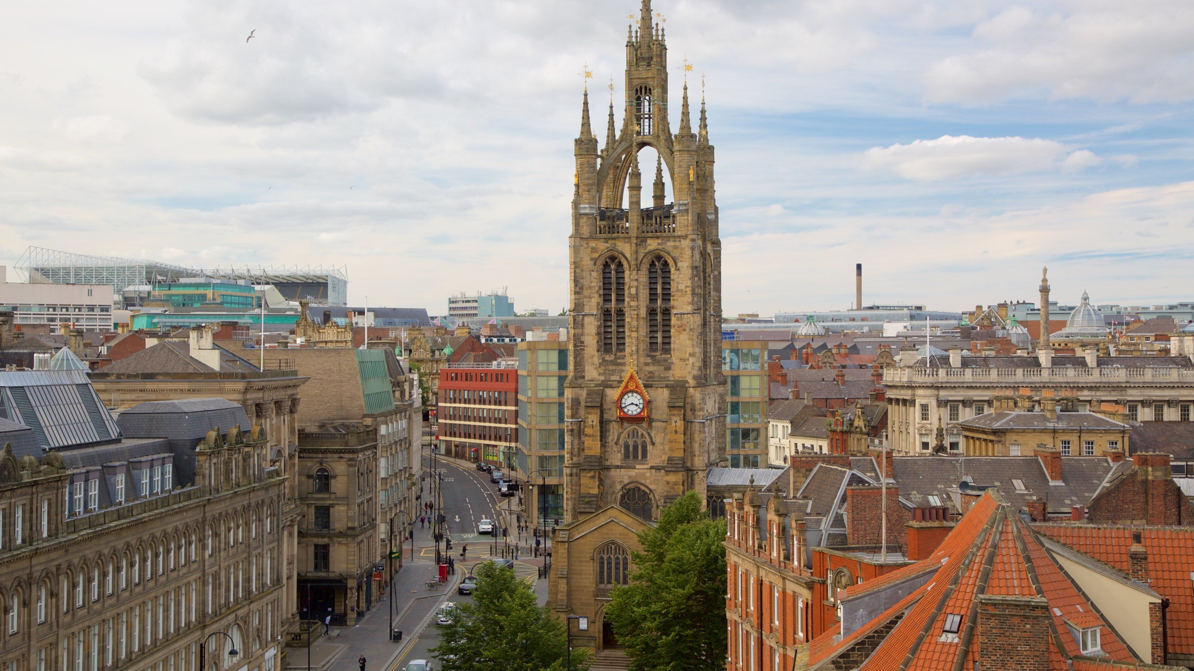 A historic church with a tall tower and a clock, surrounded by urban buildings and a modern stadium.