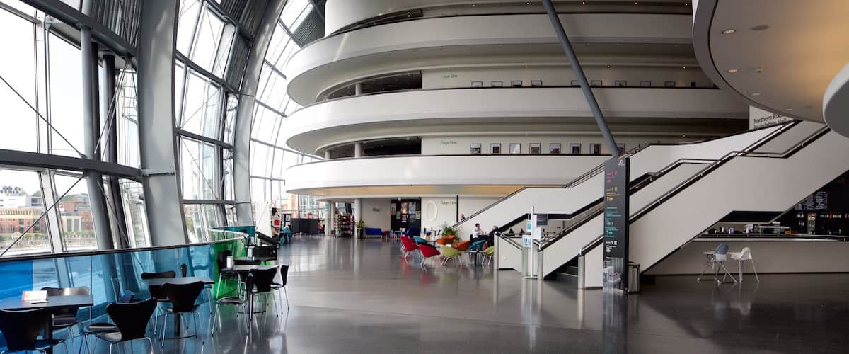 A modern indoor space with large windows, a central staircase, and seating areas.