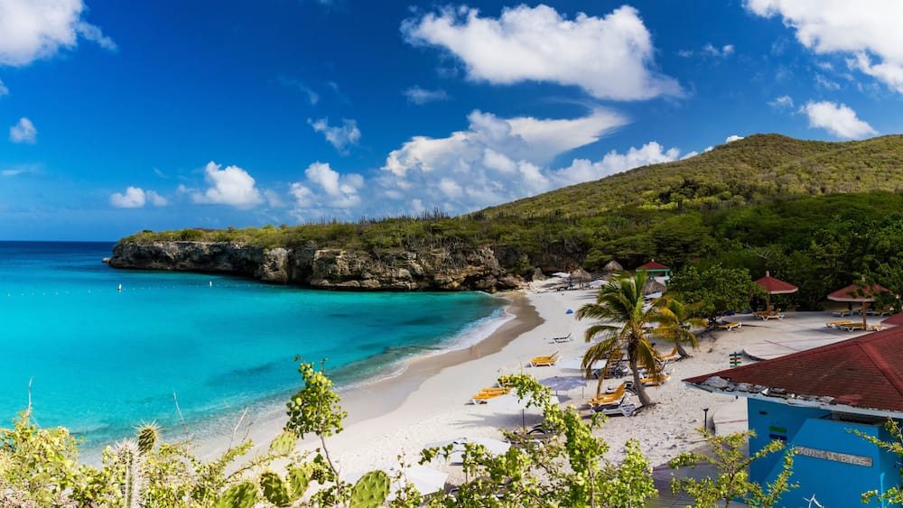 Ein Strand mit klarem türkisblauem Wasser, weißem Sand und einer felsigen Klippe. Palmen und Strandstühle sind zu sehen.