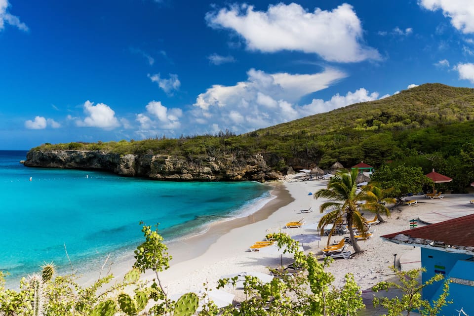 Une plage avec une eau turquoise et limpide, du sable blanc et une falaise rocheuse. On y trouve des palmiers et des parasols de plage.