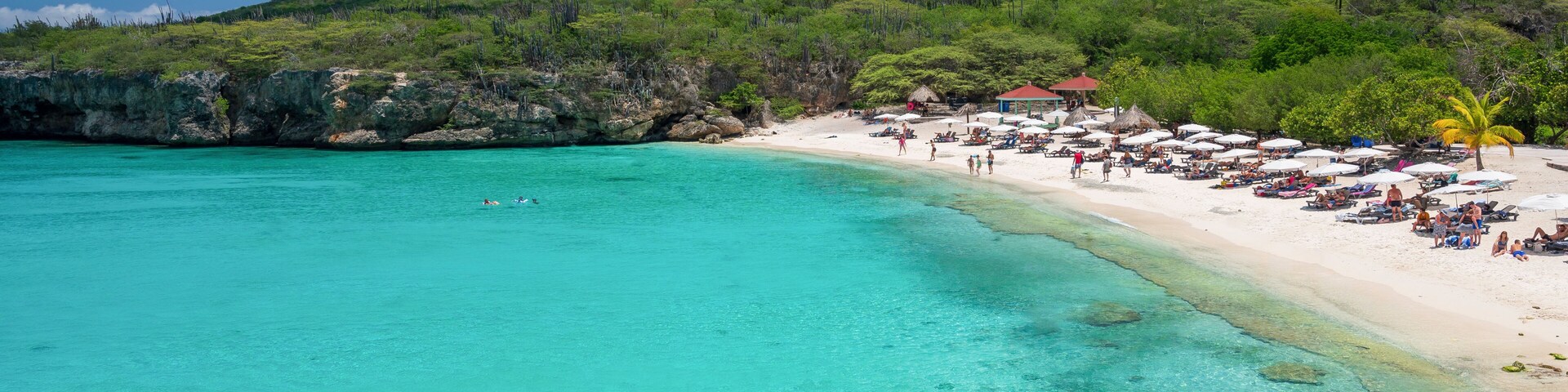 Uma praia turquesa com águas cristalinas, areal arenoso e uma colina verde ao fundo.