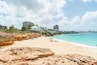 A beach with clear turquoise water, rocky foreground, and buildings in the background.