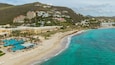A coastal resort with a turquoise beach, a swimming pool, and hillside buildings.
