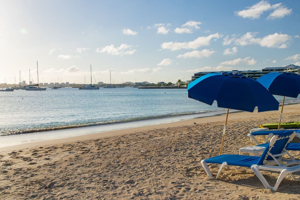 A beach with blue umbrellas and lounge chairs, a sandy shore, and sailboats in the water.