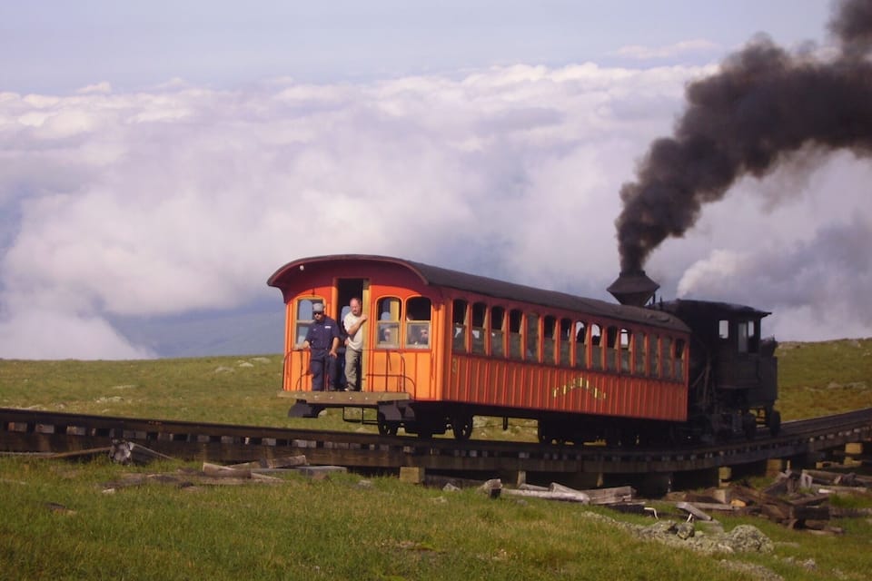 Ett ånglok med passagerare på rälsen, omgivet av gröna kullar och moln.
