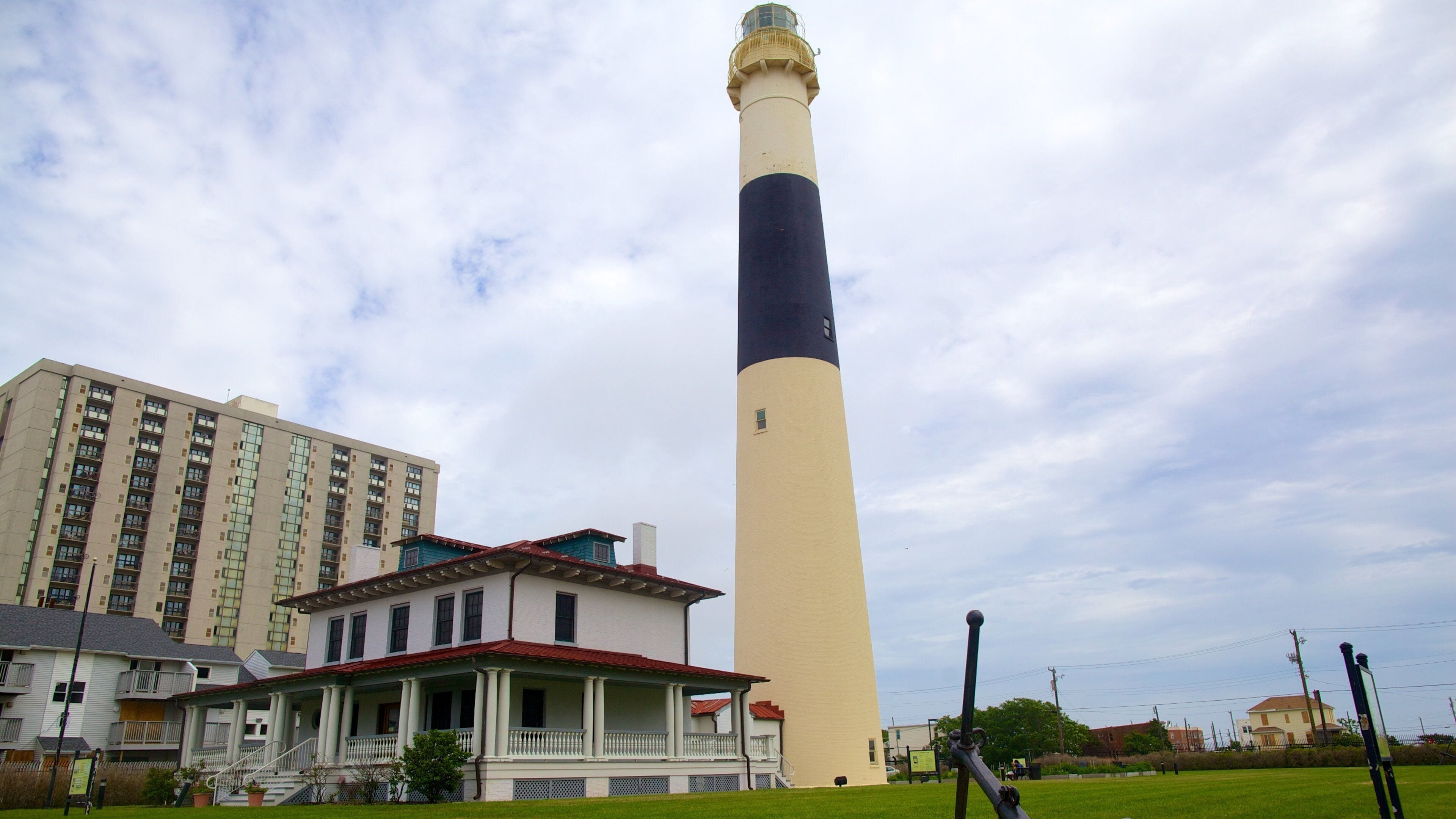 A tall lighthouse with a beige and dark blue tower, a white building with a red roof, and a grassy area with a swing set.