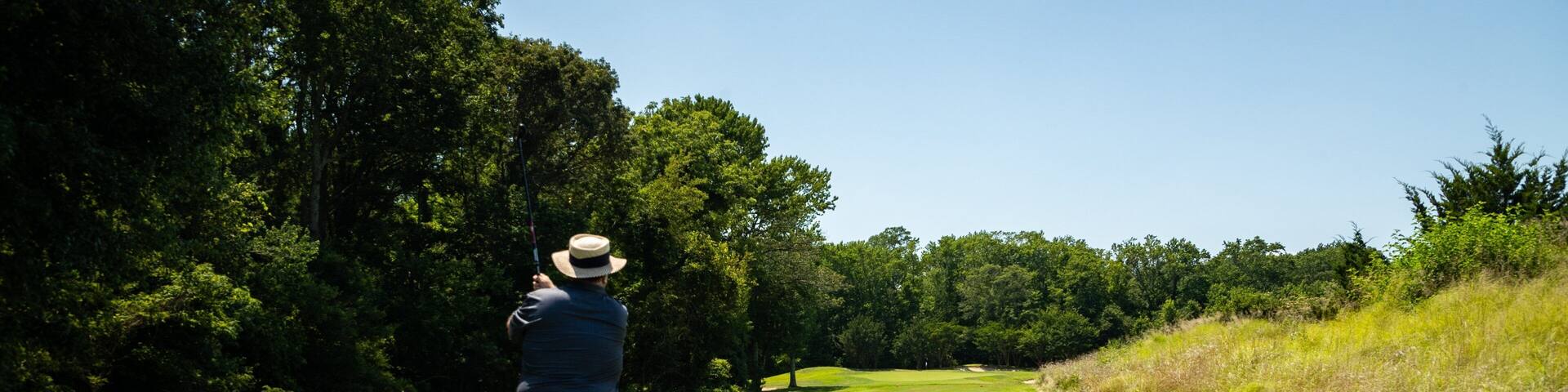 A person on a golf course with a hat, a blue shirt, and shorts, holding a golf club. There are golf balls on the grass.