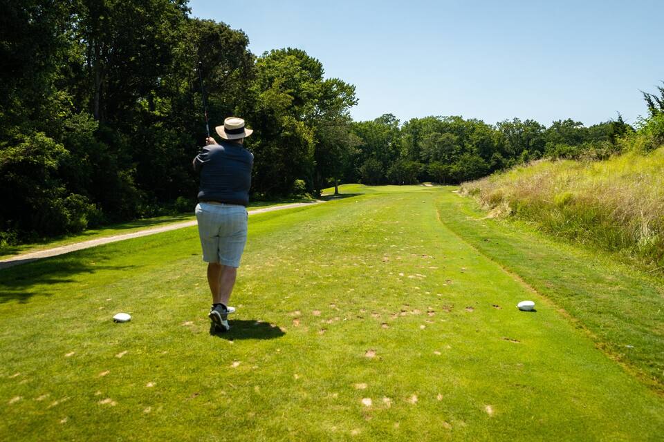 Una persona en un campo de golf, con sombrero, camisa azul y pantalones cortos, sosteniendo un palo de golf. Hay pelotas de golf sobre el césped.