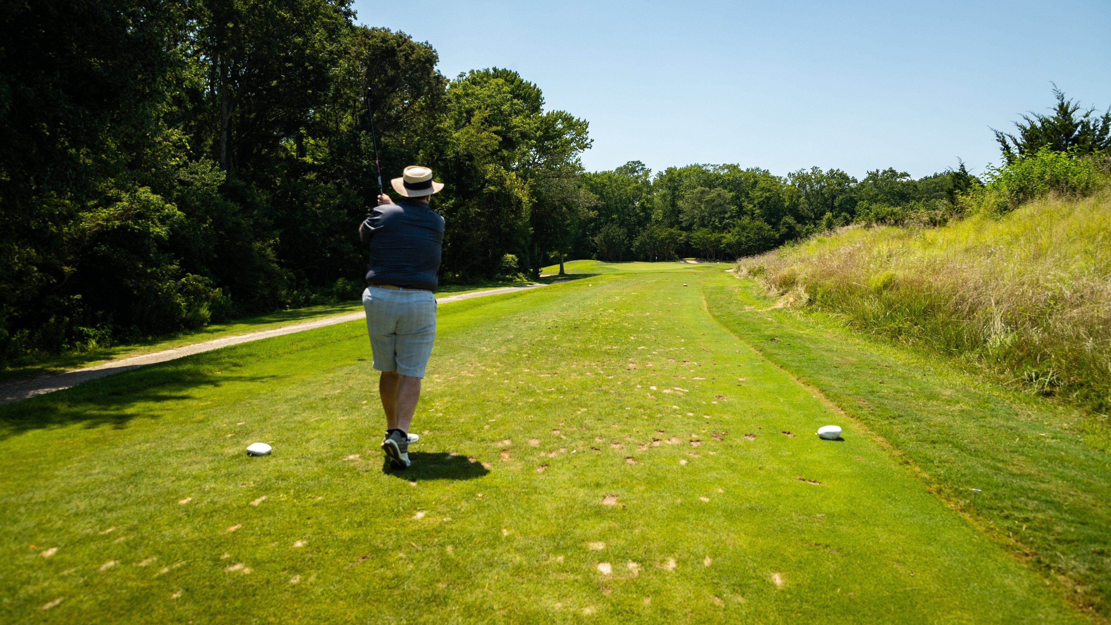 A person on a golf course with a hat, a blue shirt, and shorts, holding a golf club. There are golf balls on the grass.