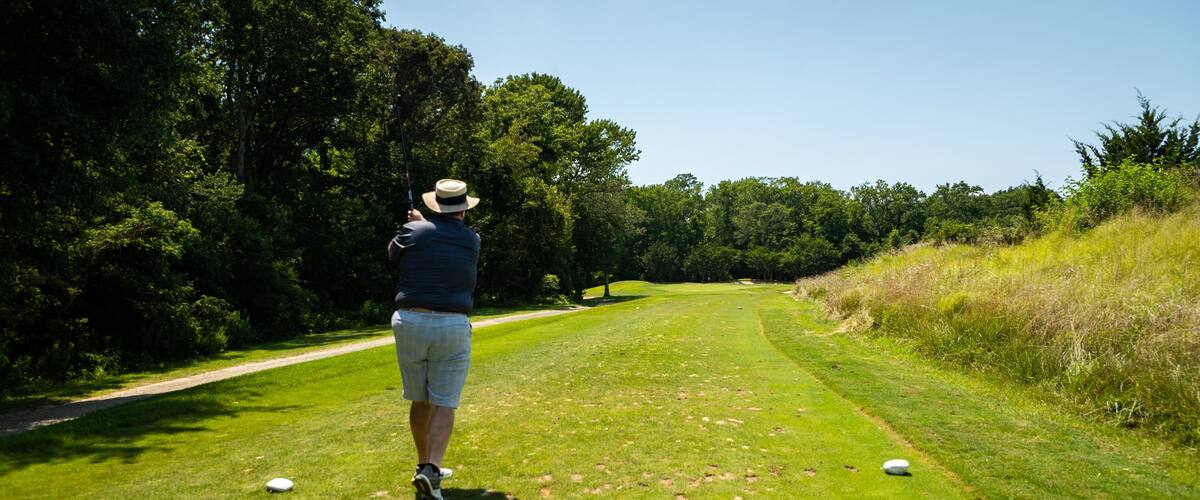A person on a golf course with a hat, a blue shirt, and shorts, holding a golf club. There are golf balls on the grass.