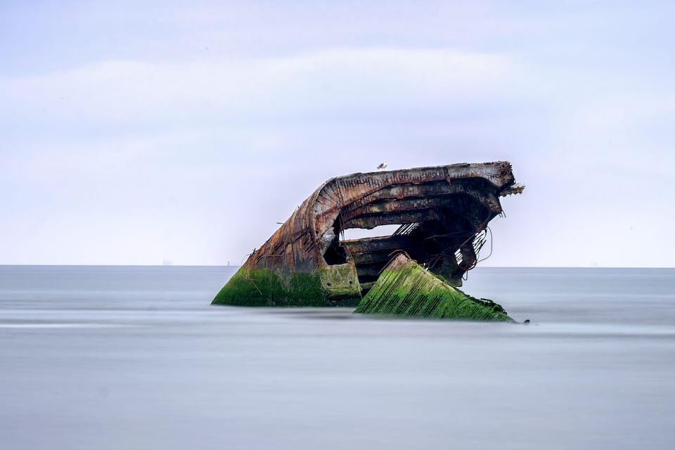 A large, rusted shipwreck partially submerged in calm water.