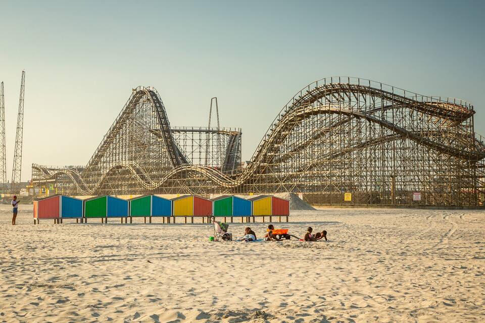 A beach with a roller coaster under construction and colorful beach huts.