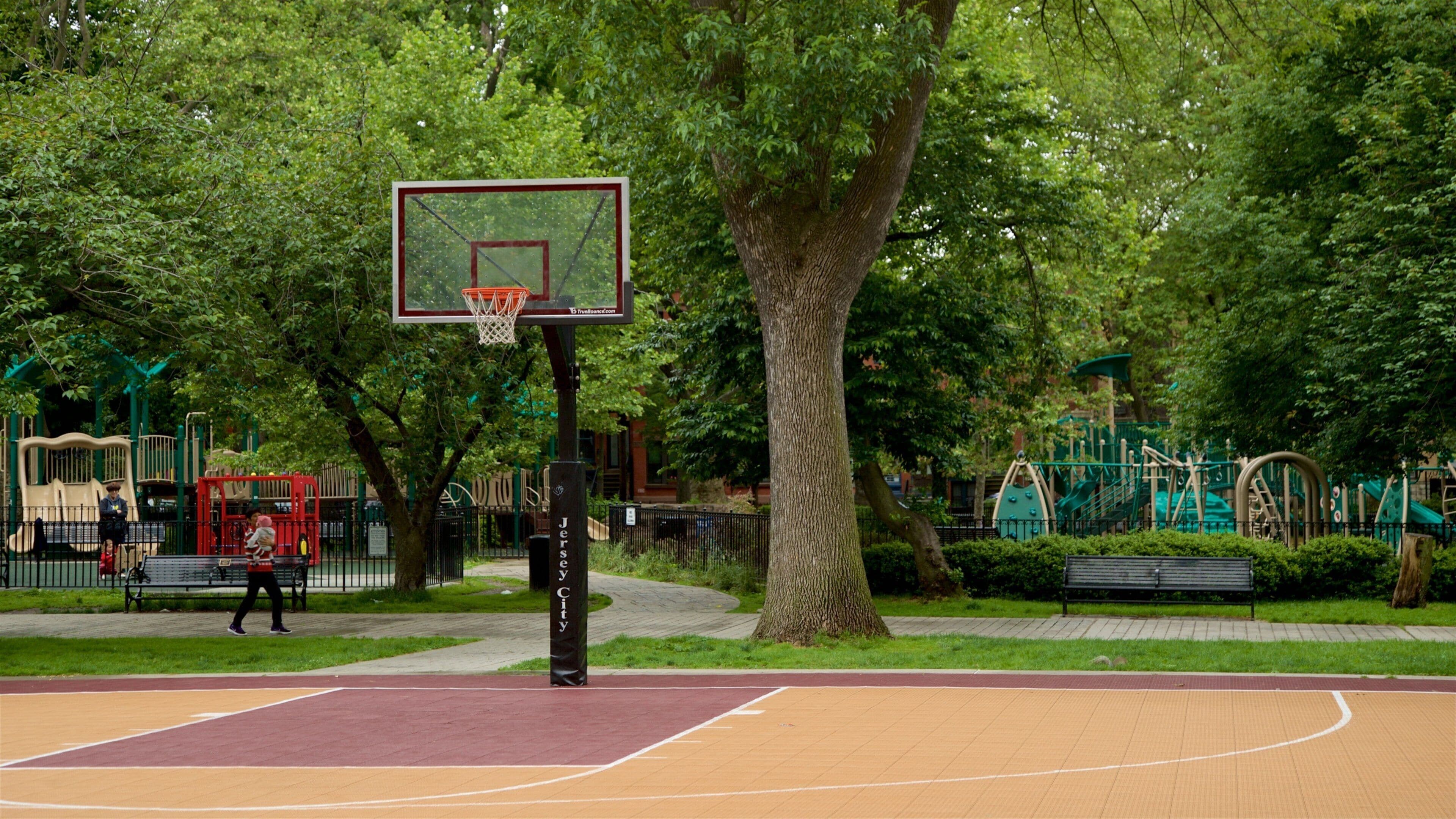 Ein Basketballplatz mit Korb, umgeben von Bäumen und Spielgeräten.