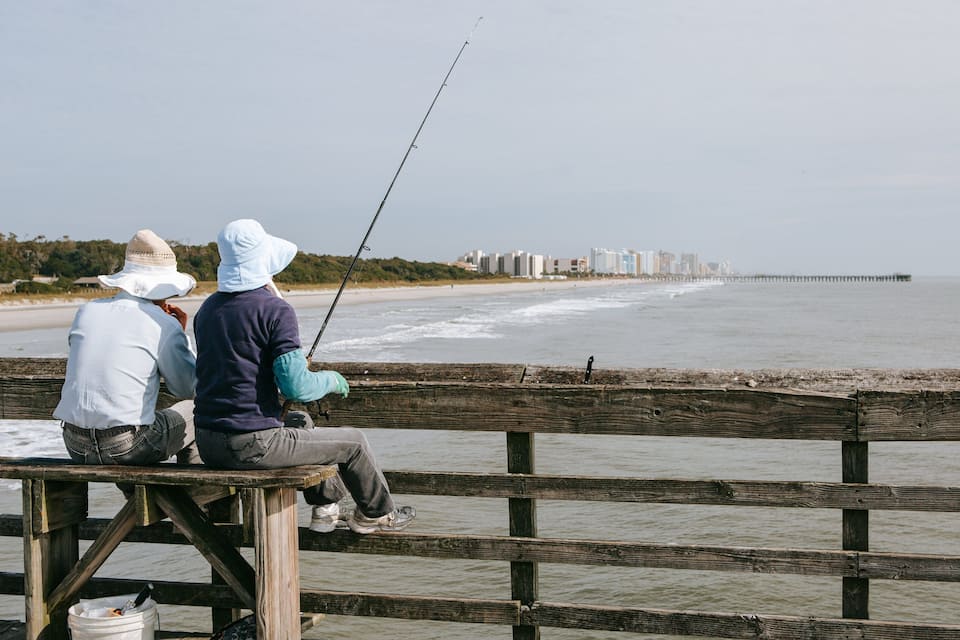 Two people fishing on a wooden pier overlooking a beach with a distant pier and buildings.