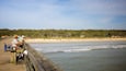 Een houten steiger met vissers, een zandstrand en een beboste heuvel op de achtergrond.