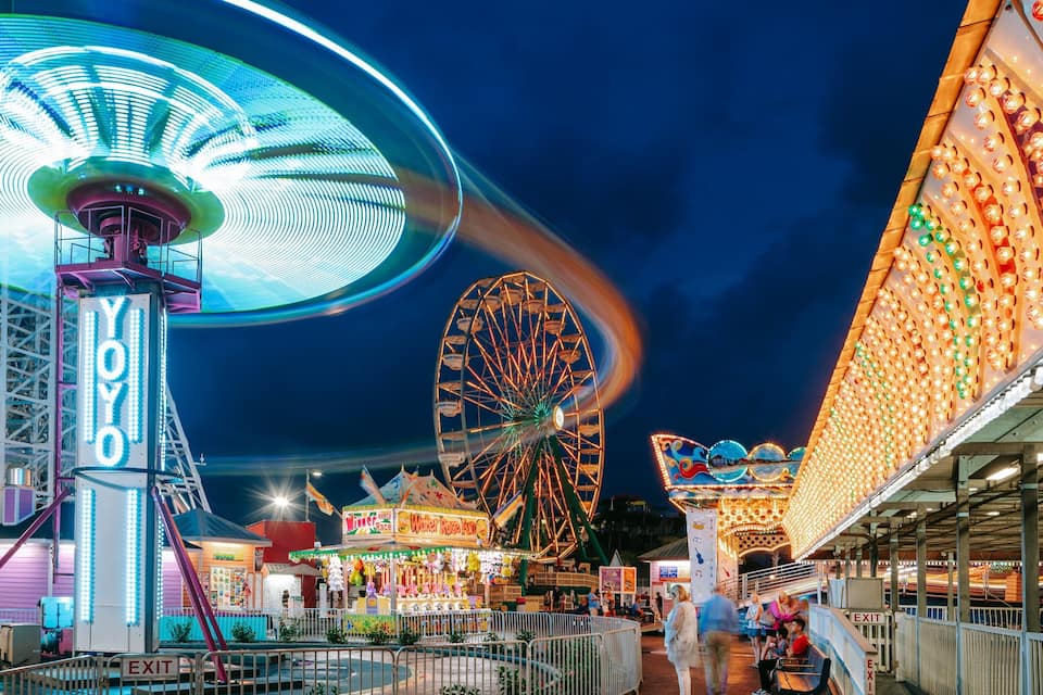 Un parque de atracciones iluminado con intensidad en la noche, con una noria, un gran juego giratorio y varios juegos de carnaval.