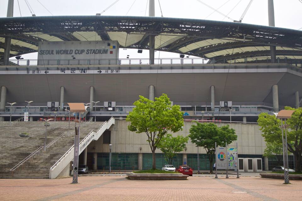 L'ingresso dello Stadio Coppa del Mondo di Seoul, con gradini che conducono all'accesso, alberi e alcune auto parcheggiate.