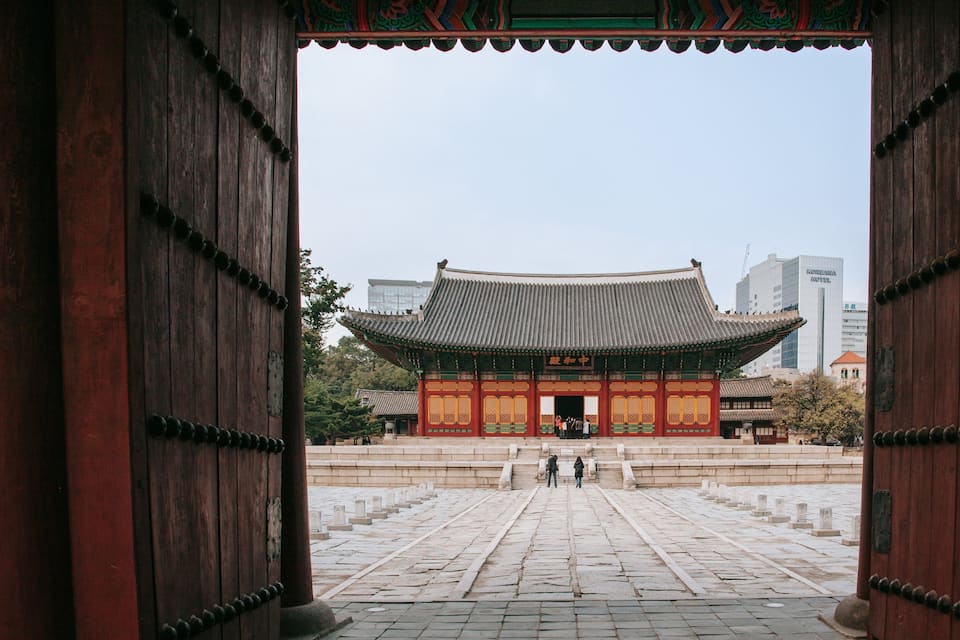 A traditional Korean gate with a tiled roof, leading to a courtyard with stone steps and a building in the background.
