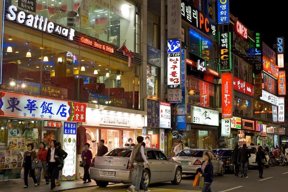A busy urban street at night with illuminated signs in multiple languages, including English and Korean.