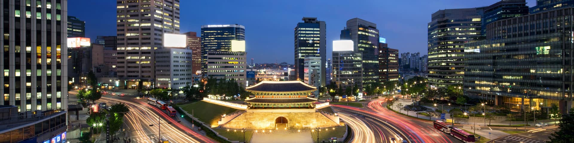 A cityscape at dusk with a traditional building illuminated in the center, surrounded by modern high-rise buildings and busy roads with light trails.