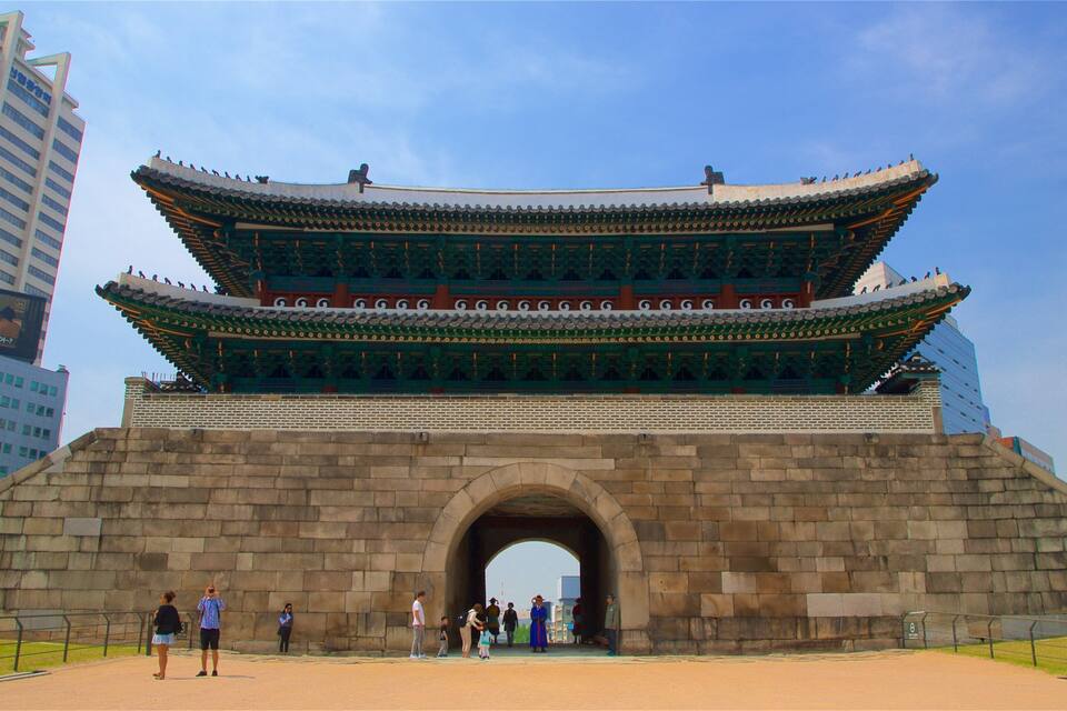 A traditional Korean gate with a curved roof and stone base, surrounded by modern buildings.