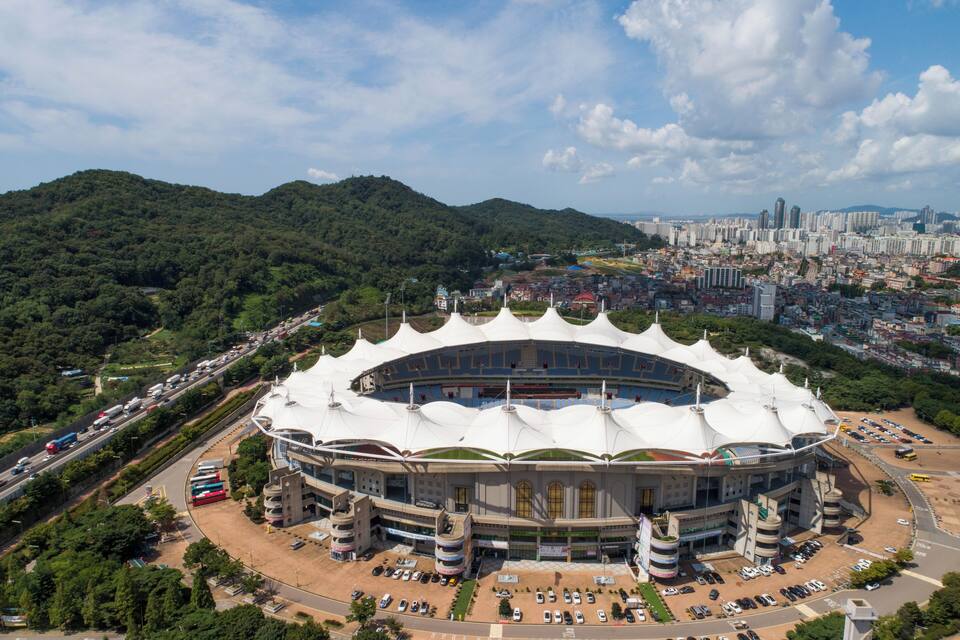 Ein großes Stadion mit weißem Dach, umgeben von einer Stadtlandschaft und grünen Hügeln.
