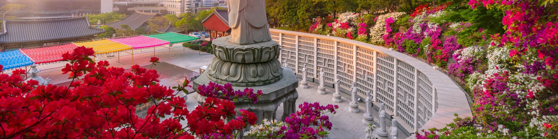 A statue with a pagoda-like roof, surrounded by vibrant flowers and a cityscape in the background.