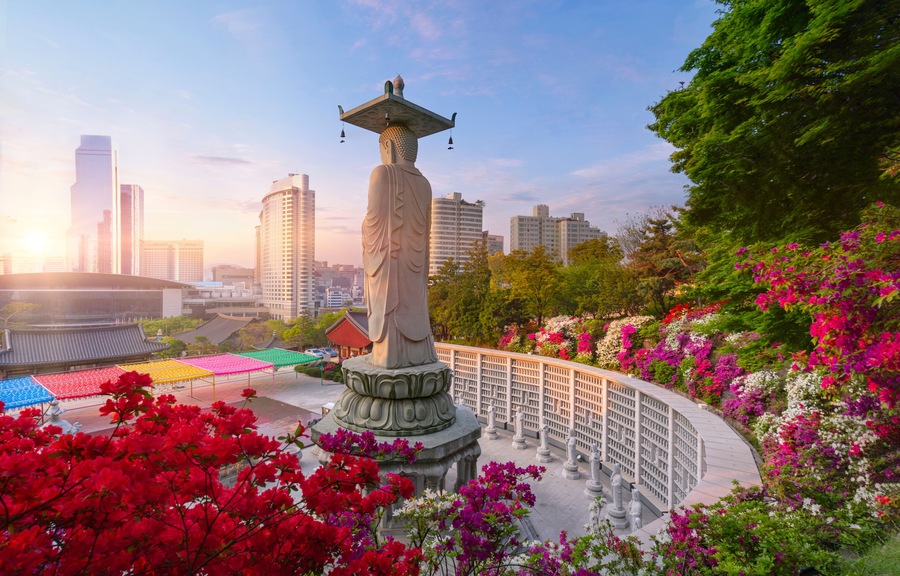 A statue with a pagoda-like roof, surrounded by vibrant flowers and a cityscape in the background.